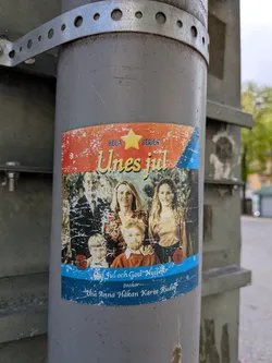 Street sticker A rectangular sticker with rounded corners, affixed to a grey utility pole, featuring a distressed, vintage aesthetic. The top section has a textured red background with white speckles, displaying a yellow star and the text 'HELA SERIEN' and 'Unes jul' in yellow script. The central part showcases a family portrait: a man, two women, and two young boys, all with a slightly faded, textured appearance. The bottom section features a distressed blue background with white speckles, containing the text 'Jul och Gott Nytt År önskar-' followed by the names 'Une Anna Håkan Karin Rudolf'. Two small red circular seals are also visible in the lower portion of the family photo.