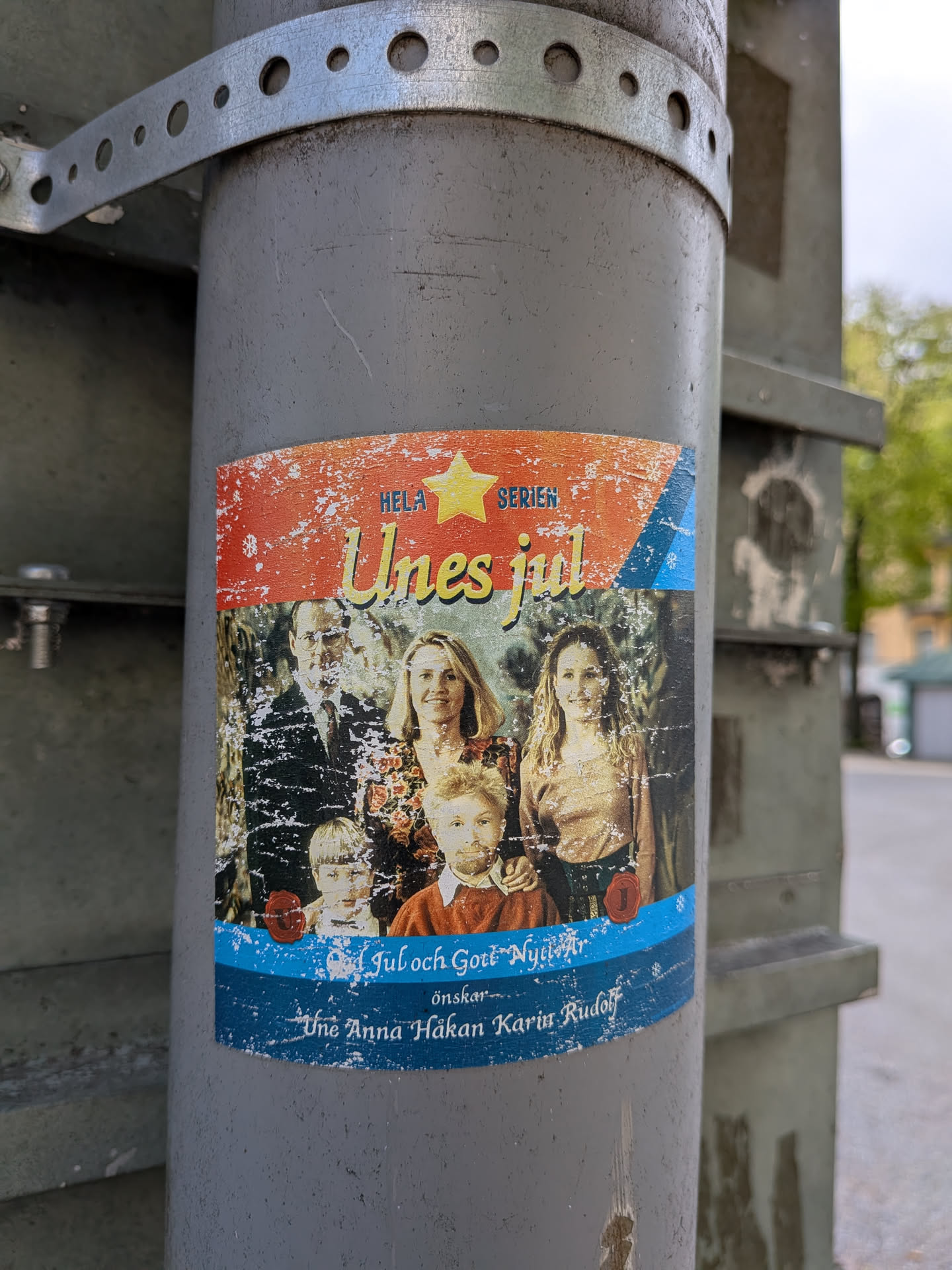 A rectangular sticker with rounded corners, affixed to a grey utility pole, featuring a distressed, vintage aesthetic. The top section has a textured red background with white speckles, displaying a yellow star and the text 'HELA SERIEN' and 'Unes jul' in yellow script. The central part showcases a family portrait: a man, two women, and two young boys, all with a slightly faded, textured appearance. The bottom section features a distressed blue background with white speckles, containing the text 'Jul och Gott Nytt År önskar-' followed by the names 'Une Anna Håkan Karin Rudolf'. Two small red circular seals are also visible in the lower portion of the family photo.