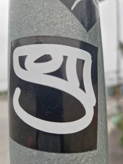 The sticker is a rectangular black vinyl with a bold, white graphic taking up most of its area. The graphic is a complex, flowing symbol with thick, rounded lines, reminiscent of calligraphic text or a highly stylized character from an Indic script, such as Devanagari. It is affixed to a textured, weathered grey metal pole, with part of another sticker visible at the top edge of the frame.