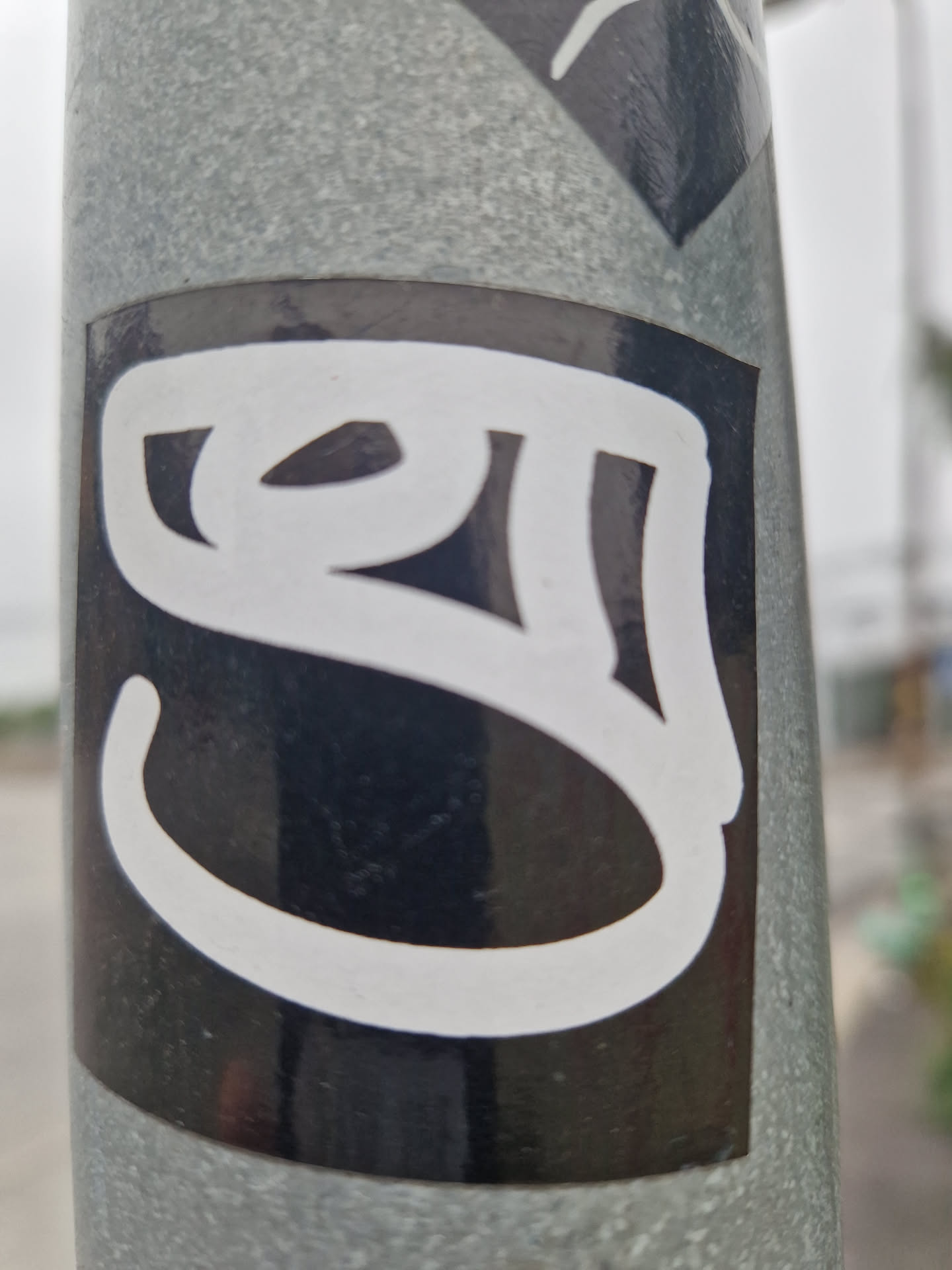 Street sticker The sticker is a rectangular black vinyl with a bold, white graphic taking up most of its area. The graphic is a complex, flowing symbol with thick, rounded lines, reminiscent of calligraphic text or a highly stylized character from an Indic script, such as Devanagari. It is affixed to a textured, weathered grey metal pole, with part of another sticker visible at the top edge of the frame.