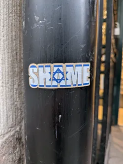 Street sticker The sticker is rectangular with slightly rounded corners, featuring the word 'SHAME' in prominent, blocky capital letters. The letters are white with a distinct blue outline. The letter 'A' in 'SHAME' is replaced by a blue Star of David symbol, which is also outlined in blue, set against the white background of the letter's form. The overall design is clean and impactful, using a simple blue and white color palette, and is adhered to a dark, scratched metal pole.
