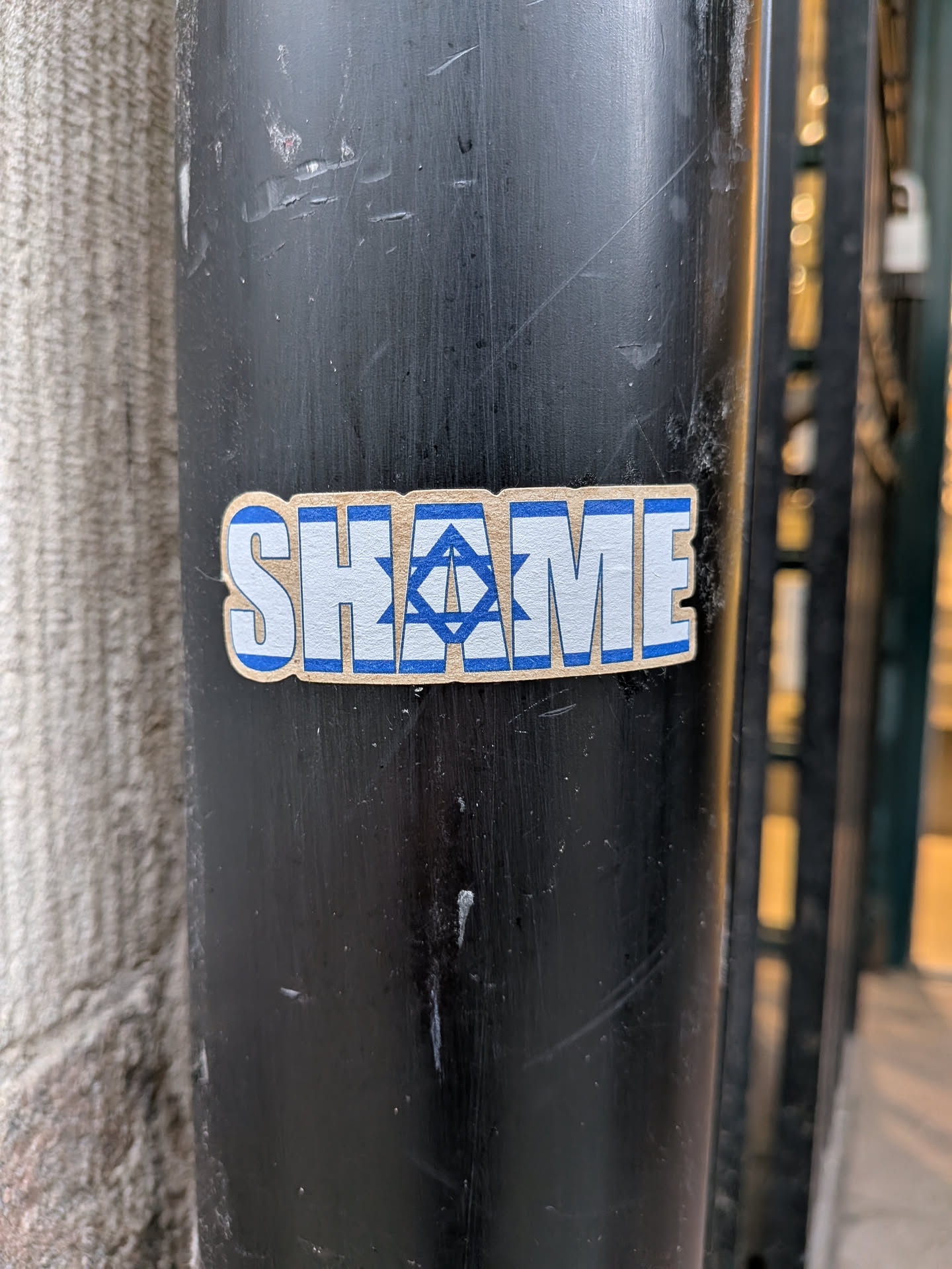 The sticker is rectangular with slightly rounded corners, featuring the word 'SHAME' in prominent, blocky capital letters. The letters are white with a distinct blue outline. The letter 'A' in 'SHAME' is replaced by a blue Star of David symbol, which is also outlined in blue, set against the white background of the letter's form. The overall design is clean and impactful, using a simple blue and white color palette, and is adhered to a dark, scratched metal pole.