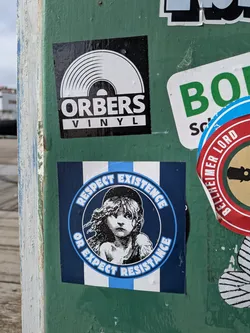 Street sticker A square sticker with a background of vertical stripes in dark blue, white, and light blue. At its center is a circular emblem with a dark blue background, featuring a black and white woodcut-style illustration of a young girl with messy hair, resembling Cosette from Les Misérables. The text 'RESPECT EXISTENCE' is arched along the top of the circle, and 'OR EXPECT RESISTANCE' is arched along the bottom, both written in a bold, light blue sans-serif font with a white outline.