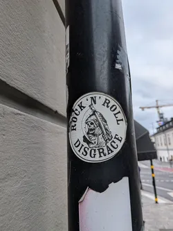 Street sticker A circular sticker with a black border and a white interior. The sticker depicts a stylized Grim Reaper in a hooded cloak, holding a scythe over its shoulder. The Grim Reaper has a grinning skull face with visible teeth. The text 'ROCK' N' ROLL' is written in an arc above the Grim Reaper, and 'DISGRACE' is written in an arc below it. The artwork is rendered in a black and white, linework style, with a slightly distressed or cracked appearance to the white background.
