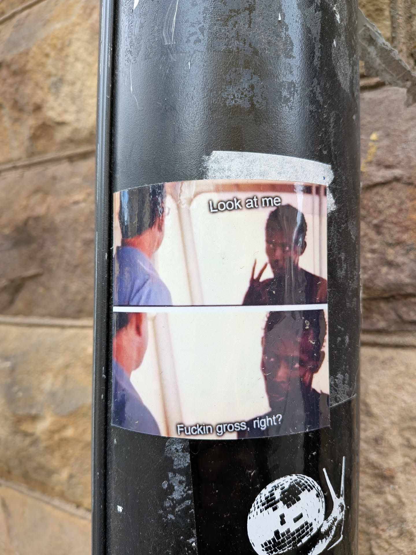 A rectangular sticker featuring a two-panel meme from the movie 'Get Out'. The top panel shows Chris Washington making a peace sign gesture, looking towards another man's back, with the text 'Look at me' above him. The bottom panel shows Chris looking directly forward with an uncomfortable expression, accompanied by the text 'Fuckin gross, right?'. The background is a light, neutral color, and the figures are depicted in naturalistic tones. The sticker is adhered to a textured black pole and shows some signs of wear and slight creasing.