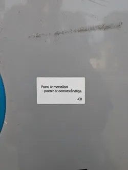 A rectangular white sticker with rounded corners on a grey metal surface. It features black Swedish text: 'Poesi är motstånd - poeter är oemotståndliga. -CB'.