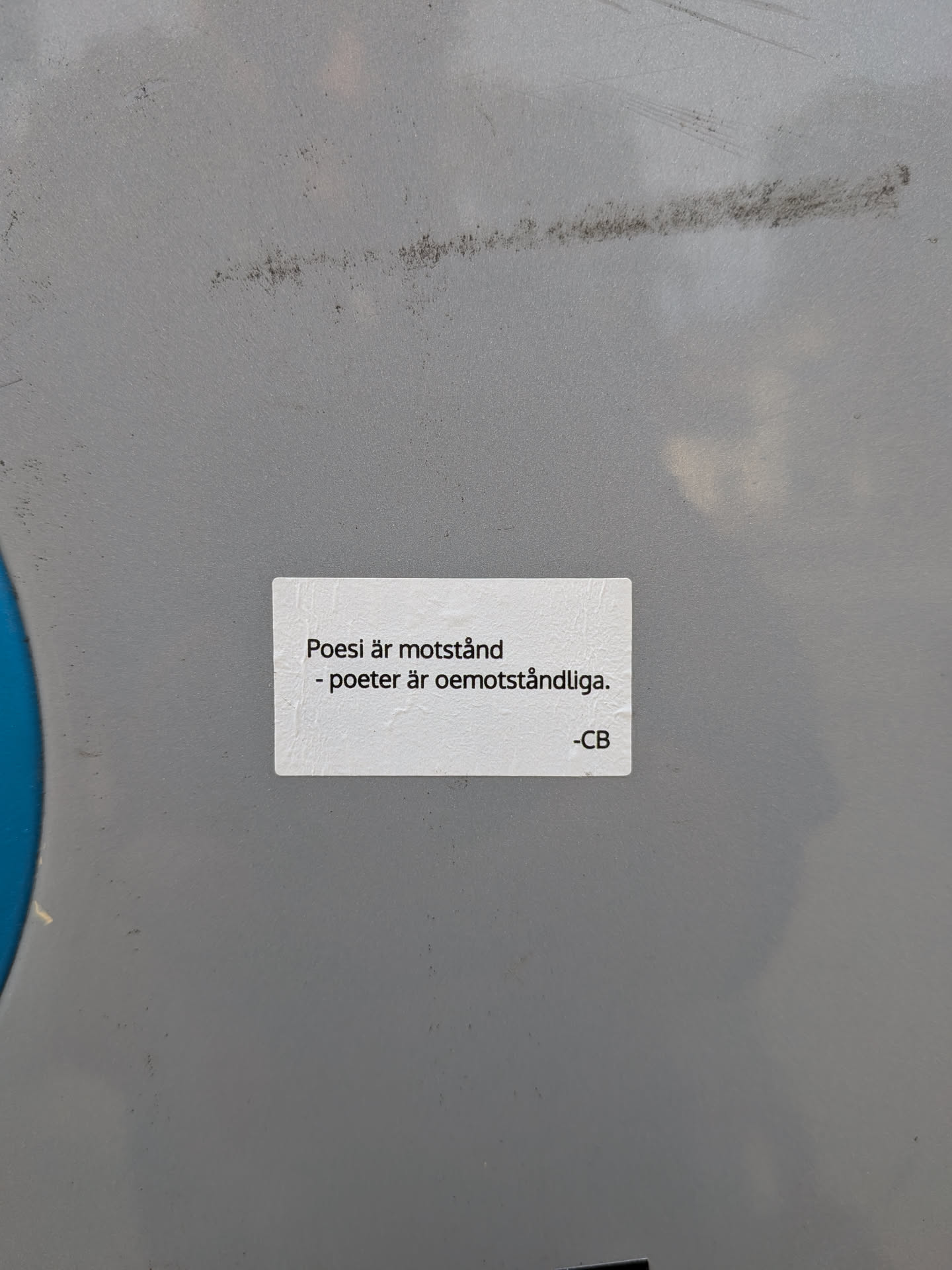 A rectangular white sticker with rounded corners on a grey metal surface. It features black Swedish text: 'Poesi är motstånd - poeter är oemotståndliga. -CB'.