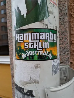 Street sticker The sticker is rectangular with a thin black outer border and a green inner border. The background features a dynamic gradient, transitioning from a fiery orange-yellow at the top, reminiscent of flares or a sunset, to a deep green at the bottom. Silhouetted against this backdrop are what appear to be a dense crowd of people, possibly at a sporting event, with bright orange light or smoke rising from within the crowd. The text "HAMMARBY" is prominently displayed at the top in a bold, white, blocky font with a black outline. Below it, "STHLM" appears in the same font. At the bottom, "Södermalm" is written in a distinctive white gothic or script-like font, also with a black outline. The overall design evokes a strong sense of local identity and football fan culture.