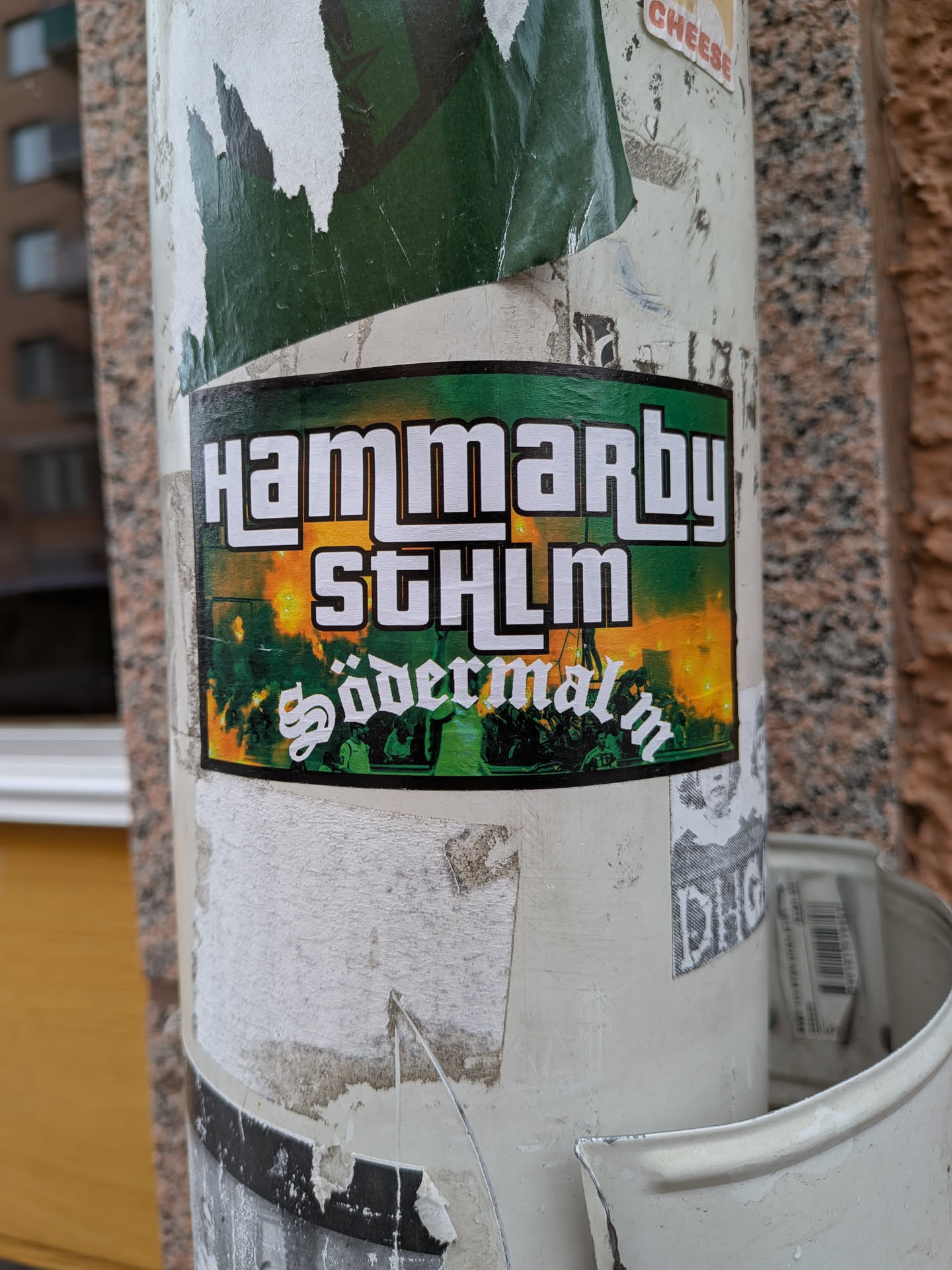 The sticker is rectangular with a thin black outer border and a green inner border. The background features a dynamic gradient, transitioning from a fiery orange-yellow at the top, reminiscent of flares or a sunset, to a deep green at the bottom. Silhouetted against this backdrop are what appear to be a dense crowd of people, possibly at a sporting event, with bright orange light or smoke rising from within the crowd. The text "HAMMARBY" is prominently displayed at the top in a bold, white, blocky font with a black outline. Below it, "STHLM" appears in the same font. At the bottom, "Södermalm" is written in a distinctive white gothic or script-like font, also with a black outline. The overall design evokes a strong sense of local identity and football fan culture.