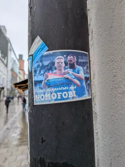 Street sticker A rectangular sticker featuring two male figures, likely football players, in light blue jerseys. The figure on the left, with blond hair, holds a flag with horizontal stripes of red, white, and light blue. The figure on the right, with dark hair and a beard, has his arm around the first. The background is a blurred image of a stadium or crowd in blue tones. Below the figures, white bold text reads 'Fotbollssupportrar mot HOMOFOBI'. The sticker is affixed to a dark, textured pole, with its top-left corner slightly peeled.