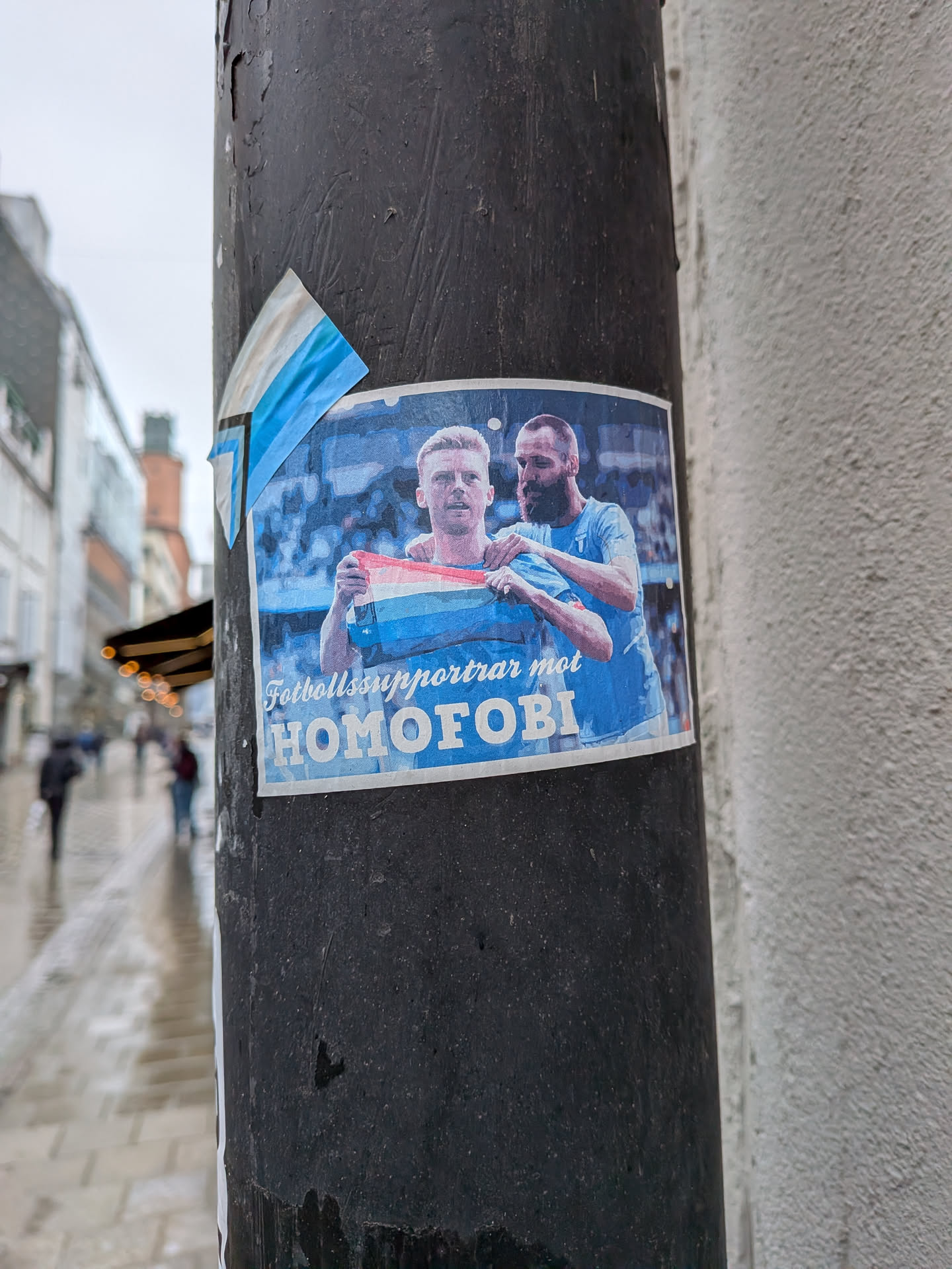 A rectangular sticker featuring two male figures, likely football players, in light blue jerseys. The figure on the left, with blond hair, holds a flag with horizontal stripes of red, white, and light blue. The figure on the right, with dark hair and a beard, has his arm around the first. The background is a blurred image of a stadium or crowd in blue tones. Below the figures, white bold text reads 'Fotbollssupportrar mot HOMOFOBI'. The sticker is affixed to a dark, textured pole, with its top-left corner slightly peeled.