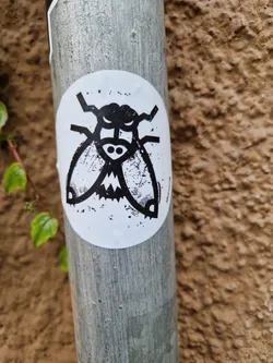 Street sticker A round sticker with a black and white illustration of a fly with angry expression. It is attached to a gray metal pole against a brown textured background. The sticker has a slightly distressed or grunge look.