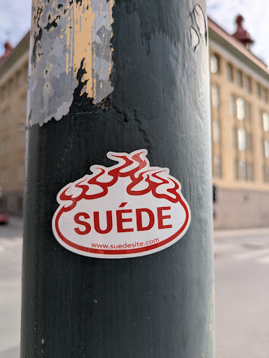 Street sticker A white sticker with a red border and red stylized flame-like shapes on top. The text 'SUÉDE' is prominently displayed in red, with the website 'www.suedesite.com' written in a smaller font below it. The sticker is affixed to a street pole with peeling paint, with a blurred building in the background.