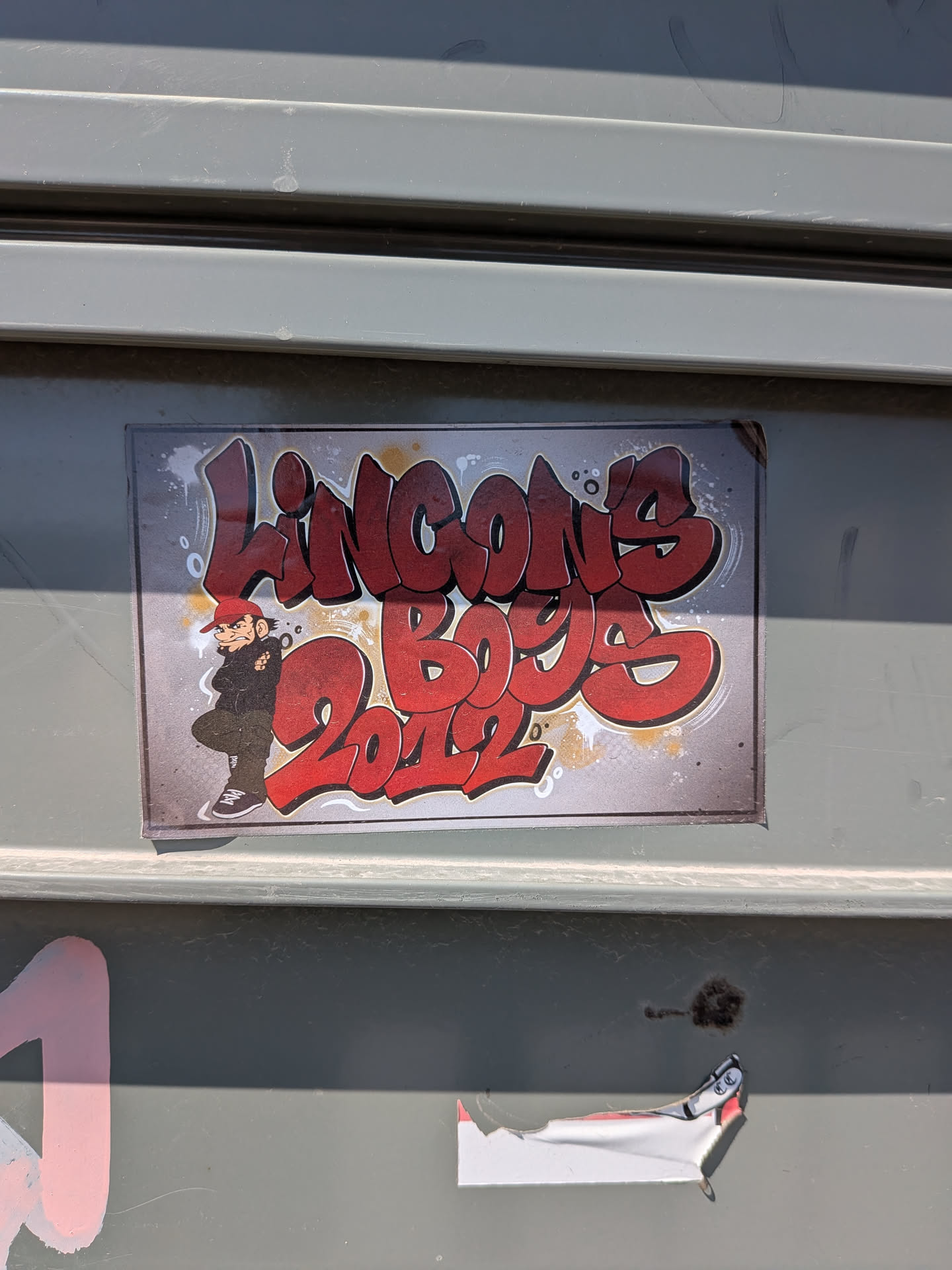 A rectangular sticker featuring red bubble-letter graffiti text 'LINCON'S BOYS 2012' on a textured grey background. A cartoon character wearing a red cap and black hoodie stands on the left side.