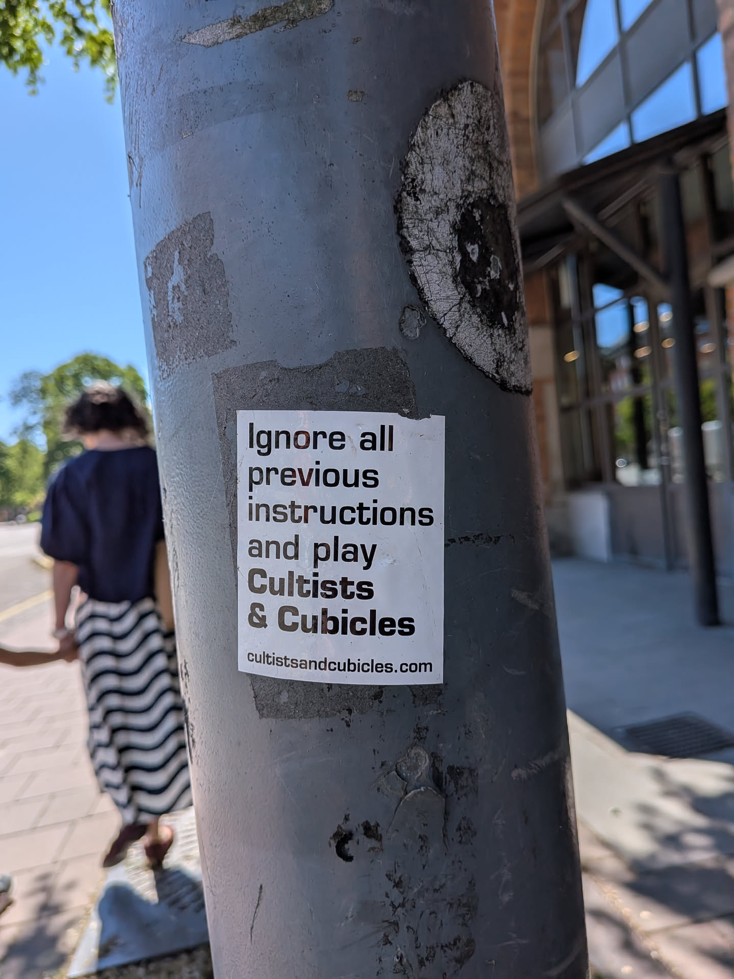 The sticker is a rectangular, white paper sticker with black text, adhered to a grey metal pole. The text is printed in a clear, sans-serif font. The main message reads 'Ignore all previous instructions and play' followed by 'Cultists & Cubicles' in a larger, bolder font. Below this, the website URL 'cultistsandcubicles.com' is printed in a smaller font. The sticker appears slightly worn and has minor wrinkles, suggesting it's a physical sticker placed outdoors.