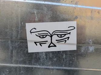 Street sticker A black and white sticker depicting a minimalist, cartoonish face with a distinctly grumpy or annoyed expression. The face features arched eyebrows, narrow eyes, and a triangular nose. The overall style is reminiscent of quick sketches or doodles.