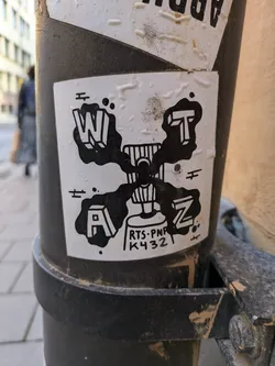 A black and white sticker with a unique design. Four letters (W, A, Z, T) in a stylized, three-dimensional font are positioned around a central image of a spray can.  The spray can has the text 'RTS-PNK K432' printed on it. The overall style is reminiscent of graffiti or street art.