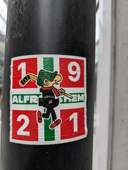 Street sticker The sticker features a whimsical cartoon character, strongly resembling Alfred E. Neuman from Mad Magazine, wearing a green checkered hat, a black outfit, and red shoes. He has a hockey stick slung over his right shoulder and is holding a smoking pipe in his left hand. The character is centered on a background designed with red, white, and green stripes, mimicking the Ikurriña (Basque flag). The top and bottom sections of the sticker feature the numbers '19' and '21' respectively, making '1921'. In the central green and white stripe, the text 'ALFR STEM' is visible.