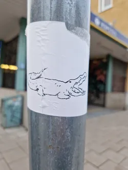 A simple line drawing of a crocodile on a white sticker affixed to a metal pole. The crocodile is depicted with its mouth open, showing its teeth. The sticker is slightly creased and shows signs of wear.
