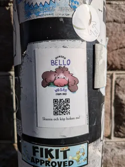 Street sticker The central sticker is rectangular with slightly rounded corners, featuring a cartoon illustration of a fluffy, brown, dog-like creature with a large pink nose and small black eyes. The creature appears to be sniffing or resting on a patch of green grass. Above the creature, in purple text, are the words 'HUNDEN BELLO'. Below the creature, in smaller purple text, it reads 'NOSAR FRAM ORD'. Below this text, a prominent black and white QR code is displayed. At the very bottom of the sticker, in black text, it says 'Skanna och köp boken nu!'. The background of the sticker is white, and it is adhered to a dark pole, surrounded by other stickers.