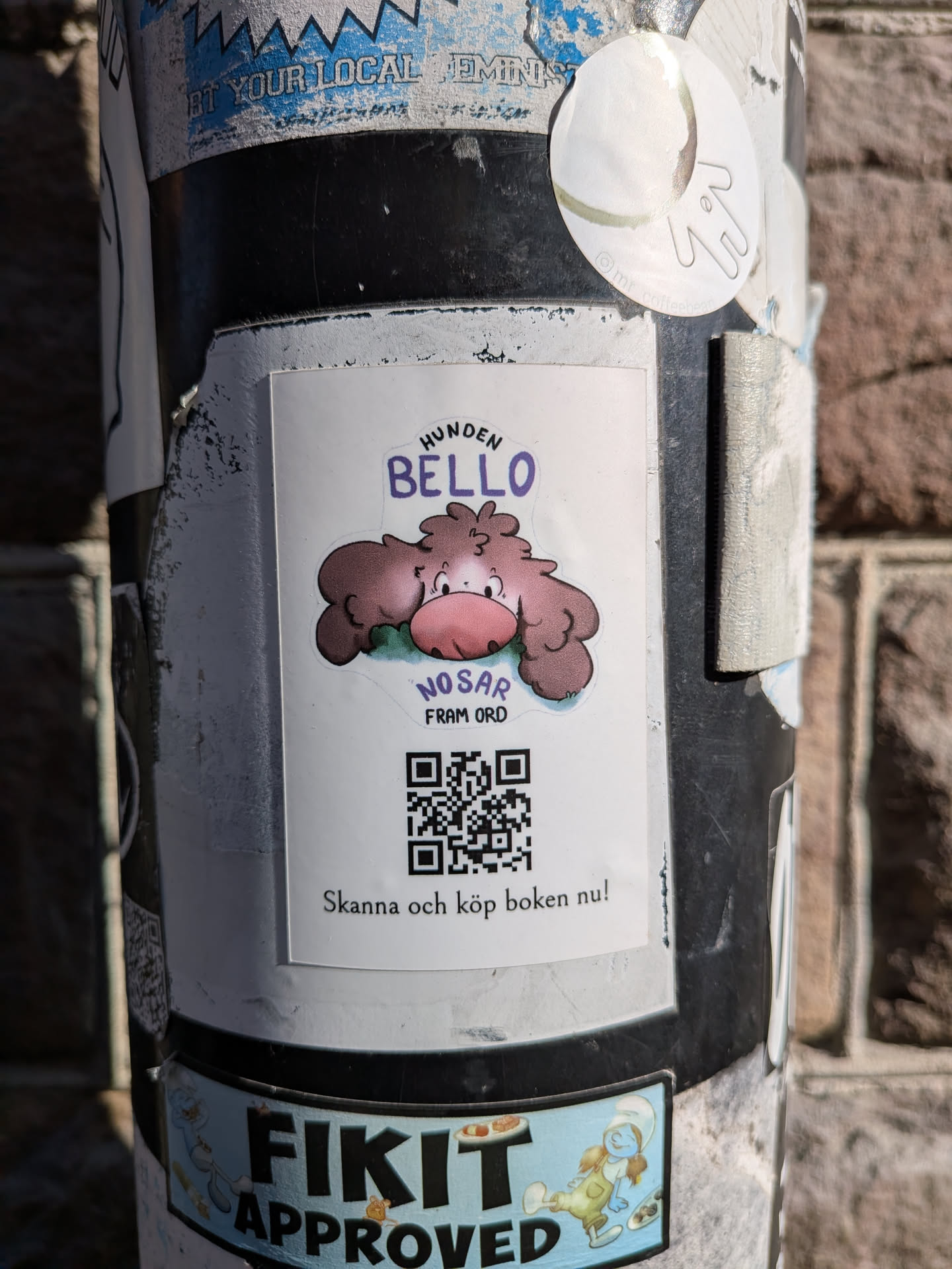 The central sticker is rectangular with slightly rounded corners, featuring a cartoon illustration of a fluffy, brown, dog-like creature with a large pink nose and small black eyes. The creature appears to be sniffing or resting on a patch of green grass. Above the creature, in purple text, are the words 'HUNDEN BELLO'. Below the creature, in smaller purple text, it reads 'NOSAR FRAM ORD'. Below this text, a prominent black and white QR code is displayed. At the very bottom of the sticker, in black text, it says 'Skanna och köp boken nu!'. The background of the sticker is white, and it is adhered to a dark pole, surrounded by other stickers.