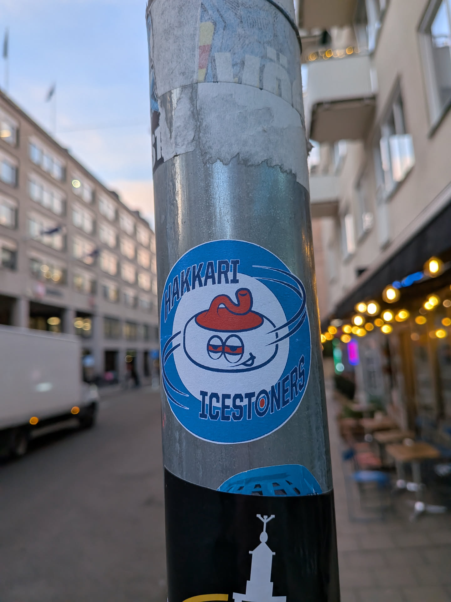 Street sticker The sticker is circular with a blue border. In the center, a cartoonish curling stone is depicted. It has large, red-rimmed eyes with pupils that appear to be looking upwards and slightly to the side, giving it a somewhat dazed or mischievous expression. A red, coiled object, possibly representing a worm or a flame, sits atop the stone like a hat. Swirling lines emanate from the stone, suggesting movement. The text 'HAKKARI' is written in an arc above the stone, and 'ICESTONERS' is written in an arc below it, both in white capital letters with a blue outline. The background of the sticker is white.