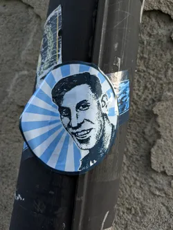 Street sticker This sticker shows a stylized portrait of a man, done in blue tones against a sunburst background. The style is reminiscent of mid-20th century graphic design.  The sticker is affixed to a dark pole outside.