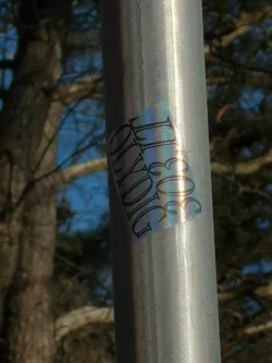 Street sticker A rectangular sticker with a holographic or iridescent background, adhered to a silver metal pole. The sticker displays a spectrum of colors, including blues, purples, and greens, which shift with the light. Dark, stylized text is printed or cut out on top of this reflective surface. The text reads '303' horizontally at the top, and 'OLYMPIG' vertically below it, rotated 90 degrees clockwise. The font is a decorative serif style, giving it an ornate appearance. The sticker is slightly angled on the pole.