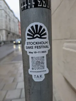 The sticker features a stylized sun design above the text "STOCKHOLM SAKE FESTIVAL" and the dates "May 10-11 2025". Below the dates is a QR code and the Instagram handle "@STOCKHOLMSAKEFESTIVAL". The sticker also includes the logo "TAK".