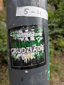 The sticker is a rectangular piece, predominantly featuring a grunge-like design in green, white, and black. The central focus is the club name "OLIMPIA" in large, vibrant green letters, stacked above "GRUDZIĄDZ" in distressed white letters with a black outline. The background is a chaotic collage of fragmented text and abstract shapes, primarily in black and white with hints of green, giving it a layered, worn appearance. In the bottom right corner, a smaller yellow-orange rectangle contains additional text. The overall aesthetic suggests a strong connection to a sports club, likely football, with a raw, fan-culture feel.