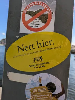 Street sticker The image shows a collection of stickers affixed to a pole. The most prominent is a yellow oval sticker with the text "Nett hier. Aber waren Sie schon mal in Baden-Württemberg?" and the Baden-Württemberg coat of arms, promoting the region with the slogan "THE LÄND". There's another sticker above, with the text "VÄLFÄRD INTE VAPEN!", showing the outline of a landmass crossed out with what seems to be bombs under it. There is also a round sticker with the text "170 JAHRE Musikverein Stephanshart".