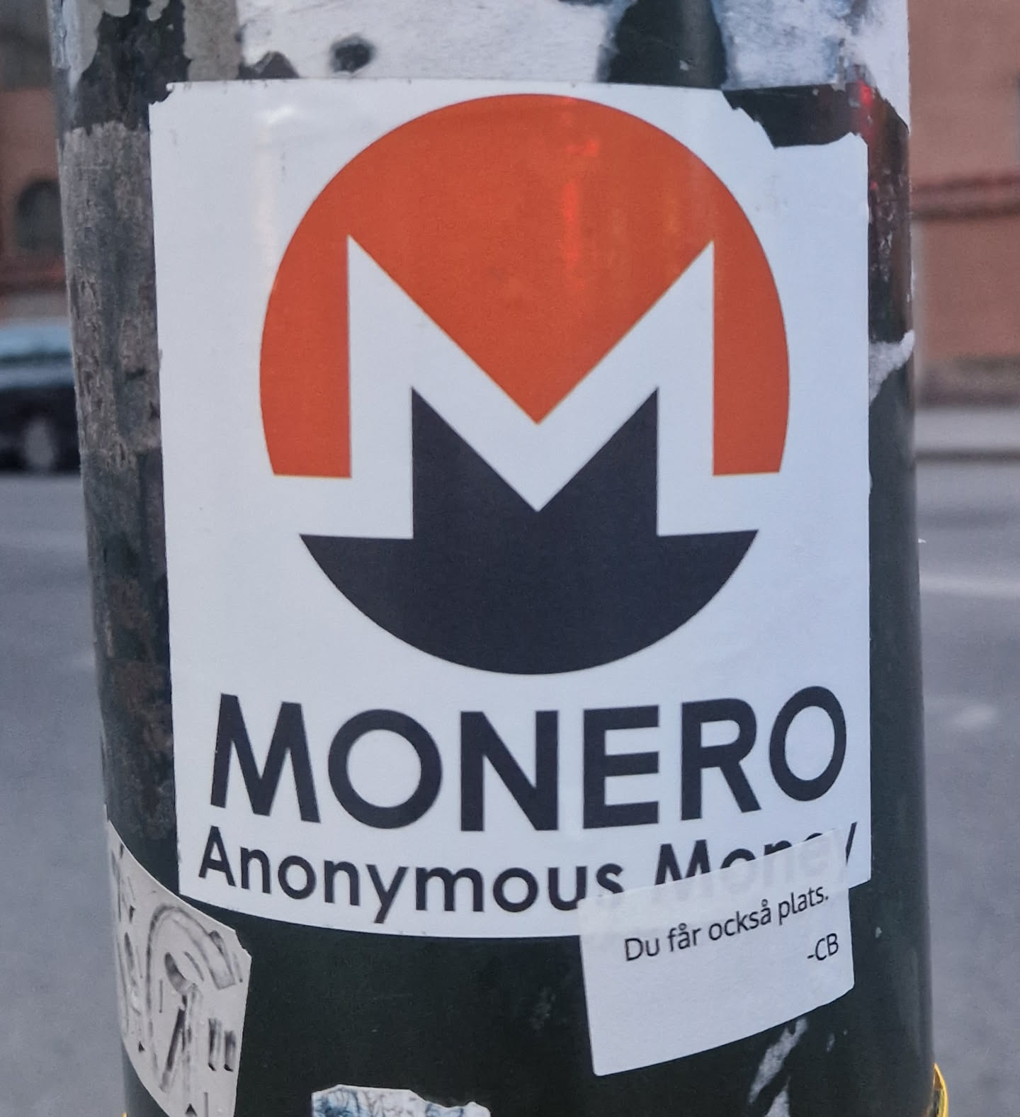 The sticker features the distinctive Monero cryptocurrency logo at its center. The logo consists of a large, vibrant orange semi-circle at the top, with a stylized black 'M' shape integrated into it, forming a unique symbol against a clean white background. Below the logo, the word 'MONERO' is printed in prominent, bold black capital letters. Beneath 'MONERO', the phrase 'Anonymous Money' is written in smaller black text. In the bottom right corner, a smaller, separate white rectangular sticker is partially visible, containing additional text. The main sticker shows slight wear and tear, with minor peeling at the edges, indicating it is affixed to a dark, textured pole.