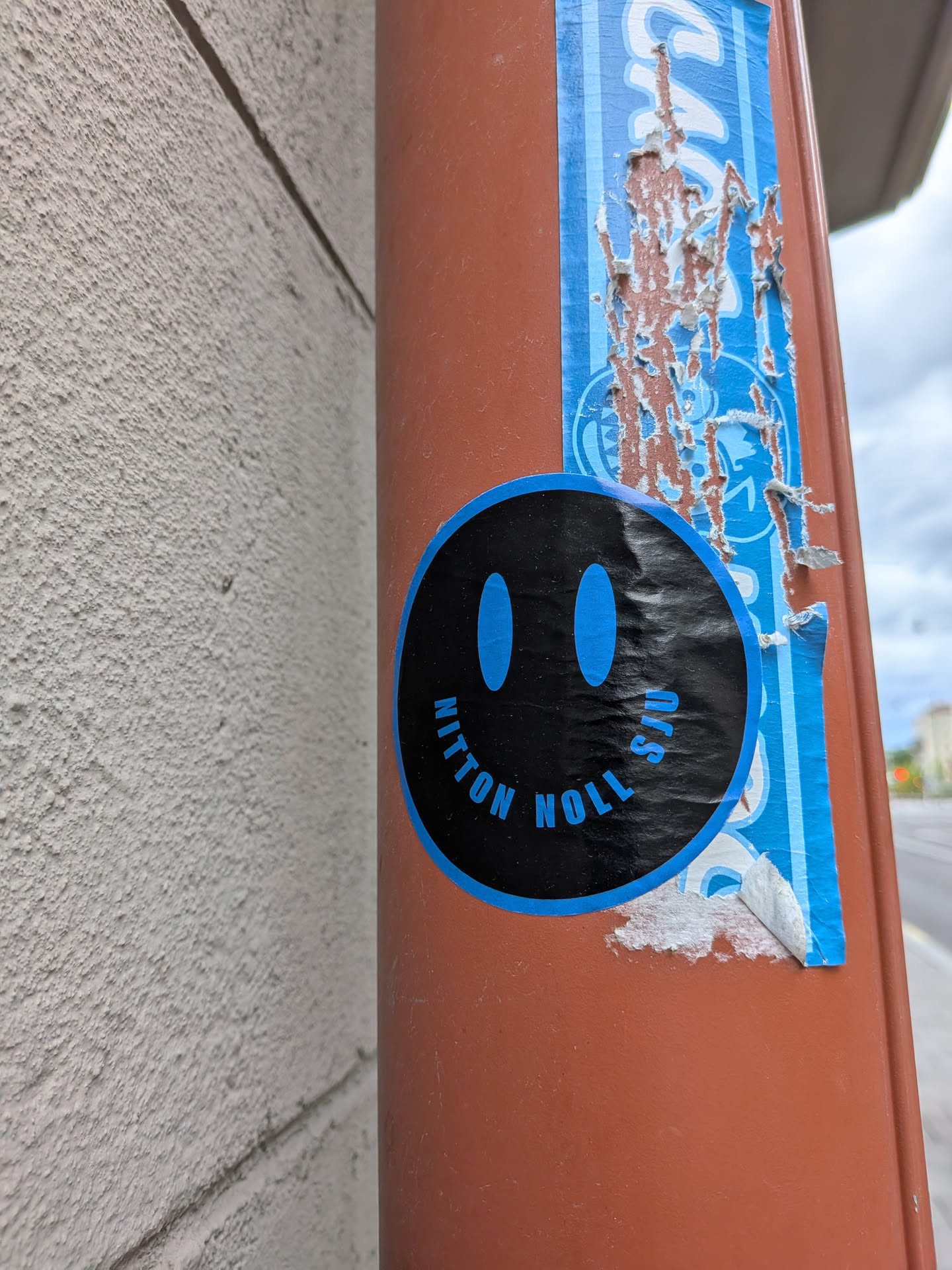 A circular sticker with a black background and blue border. It features two vertical oval eyes and the text 'NITTON NOLL SJU' curved to form a smiling mouth.
