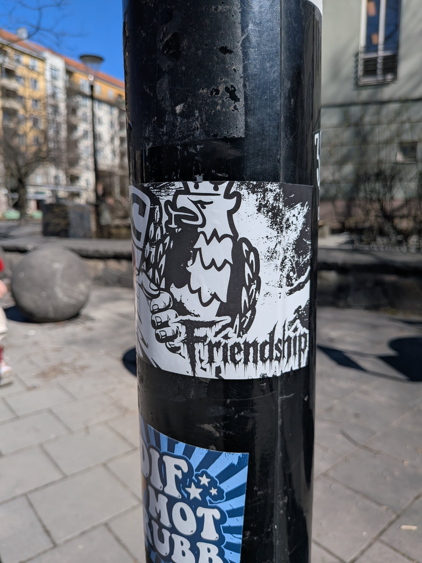 The sticker is a black and white illustration featuring a stylized, somewhat aggressive-looking creature, possibly a bird or a monster, with a prominent beak or mouth. A human-like hand, depicted with strong lines, is shown gripping the creature's neck or body. The artwork has a distressed, almost grunge or punk aesthetic, with rough black edges and splatters on a white background. Below the main illustration, the word "Friendship" is written in a gothic or heavy metal-style font. The sticker is rectangular and appears to be adhered to a textured black pole, showing some signs of wear around its edges.