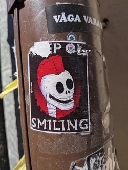 Street sticker A rectangular sticker featuring a cartoonish white skull with large black eye sockets and a wide, toothy grin. The skull sports a vibrant red punk mohawk. The background of the central image is dark, almost black, with a distressed or textured appearance. A thin white border frames the skull illustration. Above the skull, the text "KEEP ON" is visible, and below it, "SMILING" is written in a similar white, blocky font. On the left side, vertically oriented, is a small social media handle "@fntstc". The sticker itself shows signs of wear, with peeling edges and creases, indicating it's a physical sticker applied to a brown, textured pole.