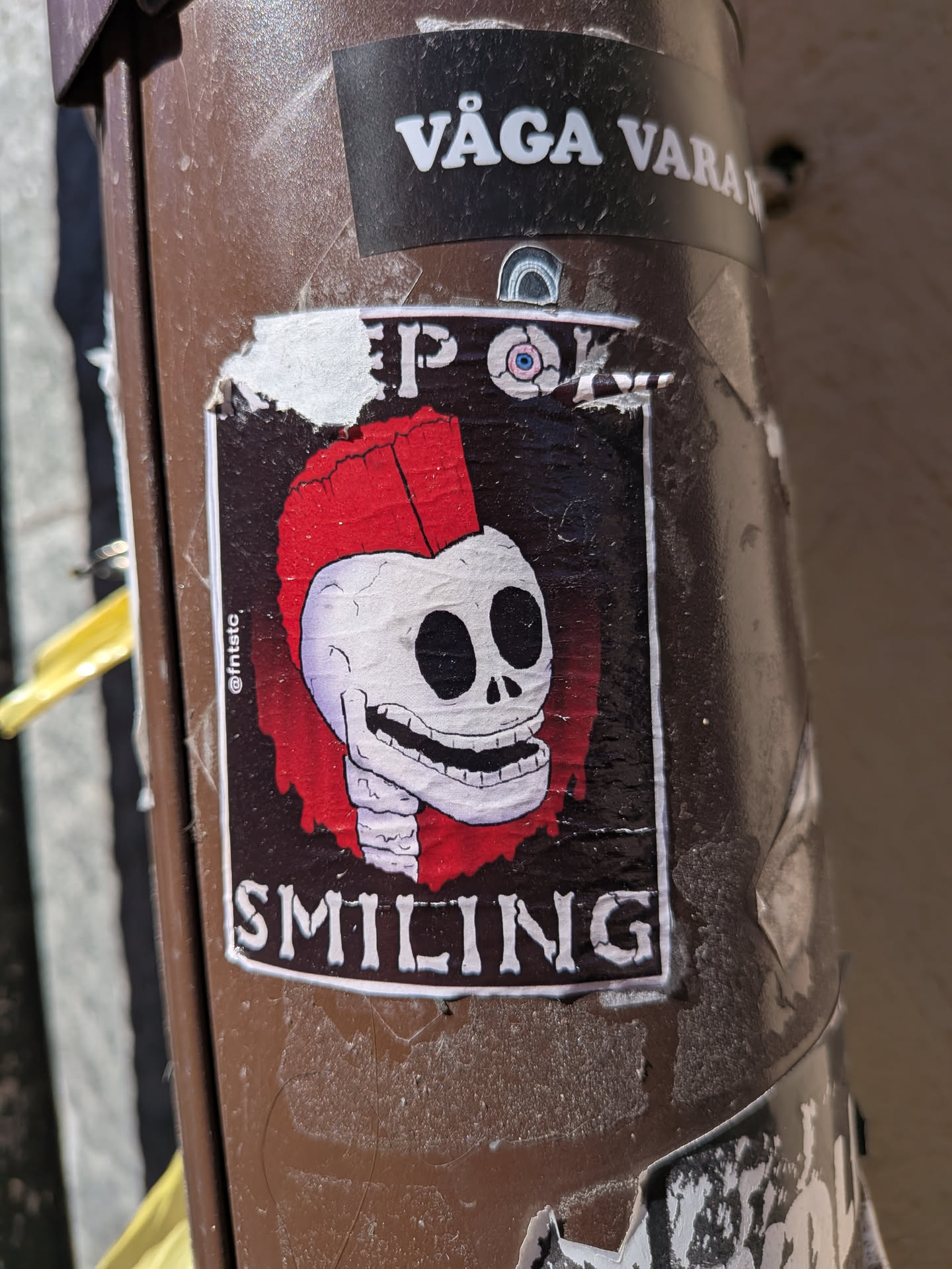 A rectangular sticker featuring a cartoonish white skull with large black eye sockets and a wide, toothy grin. The skull sports a vibrant red punk mohawk. The background of the central image is dark, almost black, with a distressed or textured appearance. A thin white border frames the skull illustration. Above the skull, the text "KEEP ON" is visible, and below it, "SMILING" is written in a similar white, blocky font. On the left side, vertically oriented, is a small social media handle "@fntstc". The sticker itself shows signs of wear, with peeling edges and creases, indicating it's a physical sticker applied to a brown, textured pole.