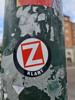 Close-up of a worn sticker affixed to a pole. The sticker features a bold red letter Z within a square, set against a white background, all inside a black circle. Below the square, the word "KLART" is clearly visible.  The sticker shows signs of age and wear, with some peeling and damage. The background shows remnants of other stickers.