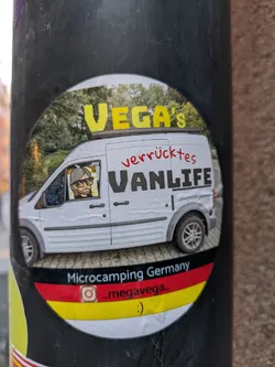 Street sticker A round sticker featuring an illustration of a white van with a person inside. The text on the sticker says "VEGA's verrücktes VANLIFE" and "Microcamping Germany". There's an Instagram handle and a smiley face at the bottom. The sticker uses the colors of the German flag.