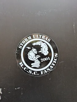 Street sticker A circular, monochromatic sticker featuring a white design on a dark, likely black or very dark grey, background. The outer ring of the sticker contains text: 'STORM ULTRAS' is arched across the top, and 'R.C.S.C. FANATICS' is arched across the bottom, both in a bold, white, serif-style font. The inner circle showcases a stylized, aggressive-looking figure in profile, facing right. The figure appears to be a hunched character, possibly wearing a helmet or hood with a pointed top, and is depicted exhaling a large, swirling cloud or puff of smoke. To the right of the figure and below the smoke, the year '2001' is prominently displayed in white. The sticker has a slightly worn appearance with minor scuffs and reflections, indicating it's a physical decal on a surface.