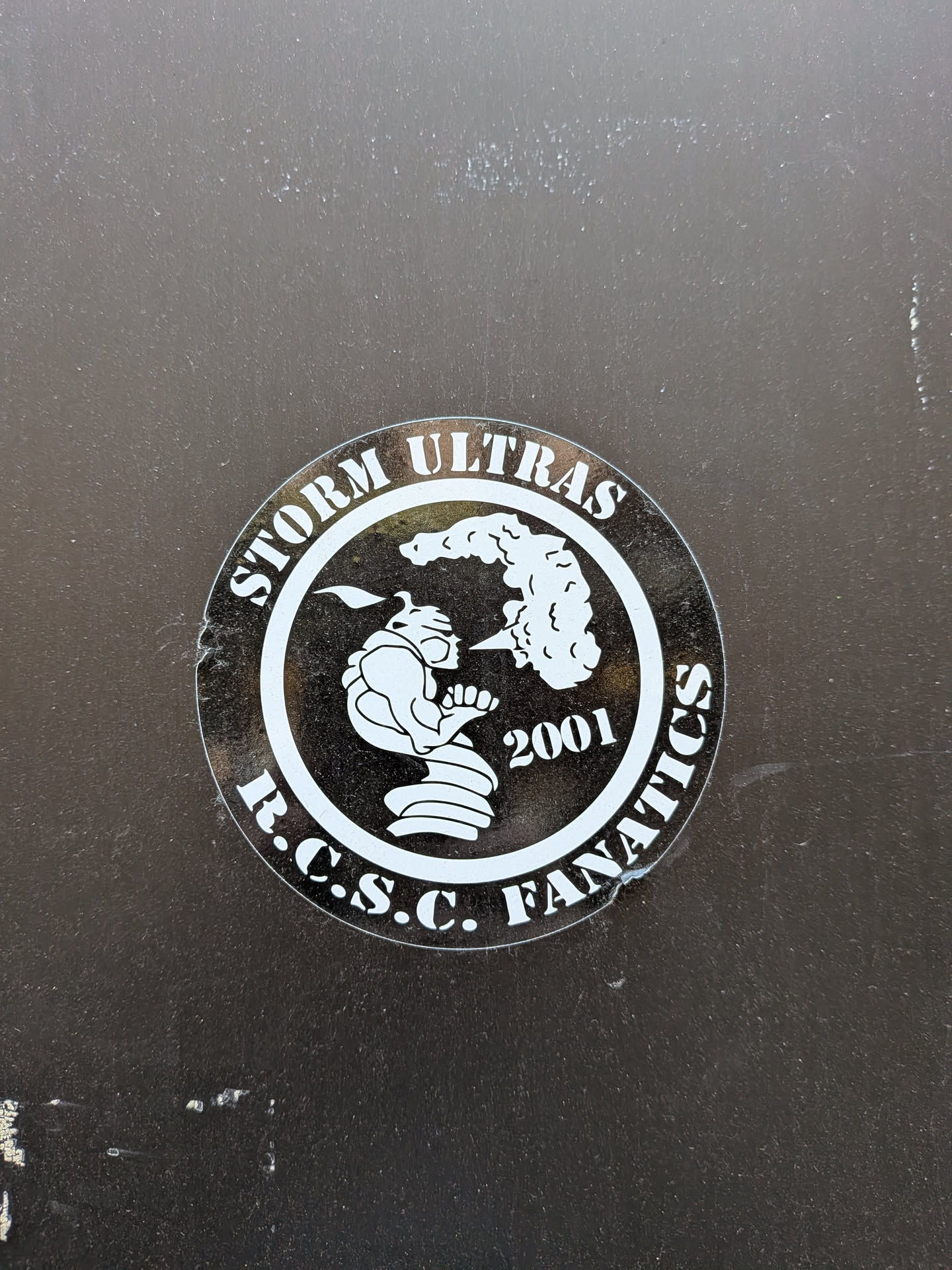 A circular, monochromatic sticker featuring a white design on a dark, likely black or very dark grey, background. The outer ring of the sticker contains text: 'STORM ULTRAS' is arched across the top, and 'R.C.S.C. FANATICS' is arched across the bottom, both in a bold, white, serif-style font. The inner circle showcases a stylized, aggressive-looking figure in profile, facing right. The figure appears to be a hunched character, possibly wearing a helmet or hood with a pointed top, and is depicted exhaling a large, swirling cloud or puff of smoke. To the right of the figure and below the smoke, the year '2001' is prominently displayed in white. The sticker has a slightly worn appearance with minor scuffs and reflections, indicating it's a physical decal on a surface.