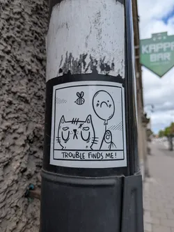 Street sticker A rectangular, black and white cartoon sticker featuring a grumpy-looking cat with closed eyes and a downturned mouth. Above the cat, a small bee is depicted in flight. To the right of the cat, a balloon with a sad face floats, and below it, a small, round creature with a paw-like hand looks upwards. The entire scene is framed by a thick black border, and below the illustration, the text 'TROUBLE FINDS ME!' is written in a black box with white lettering.