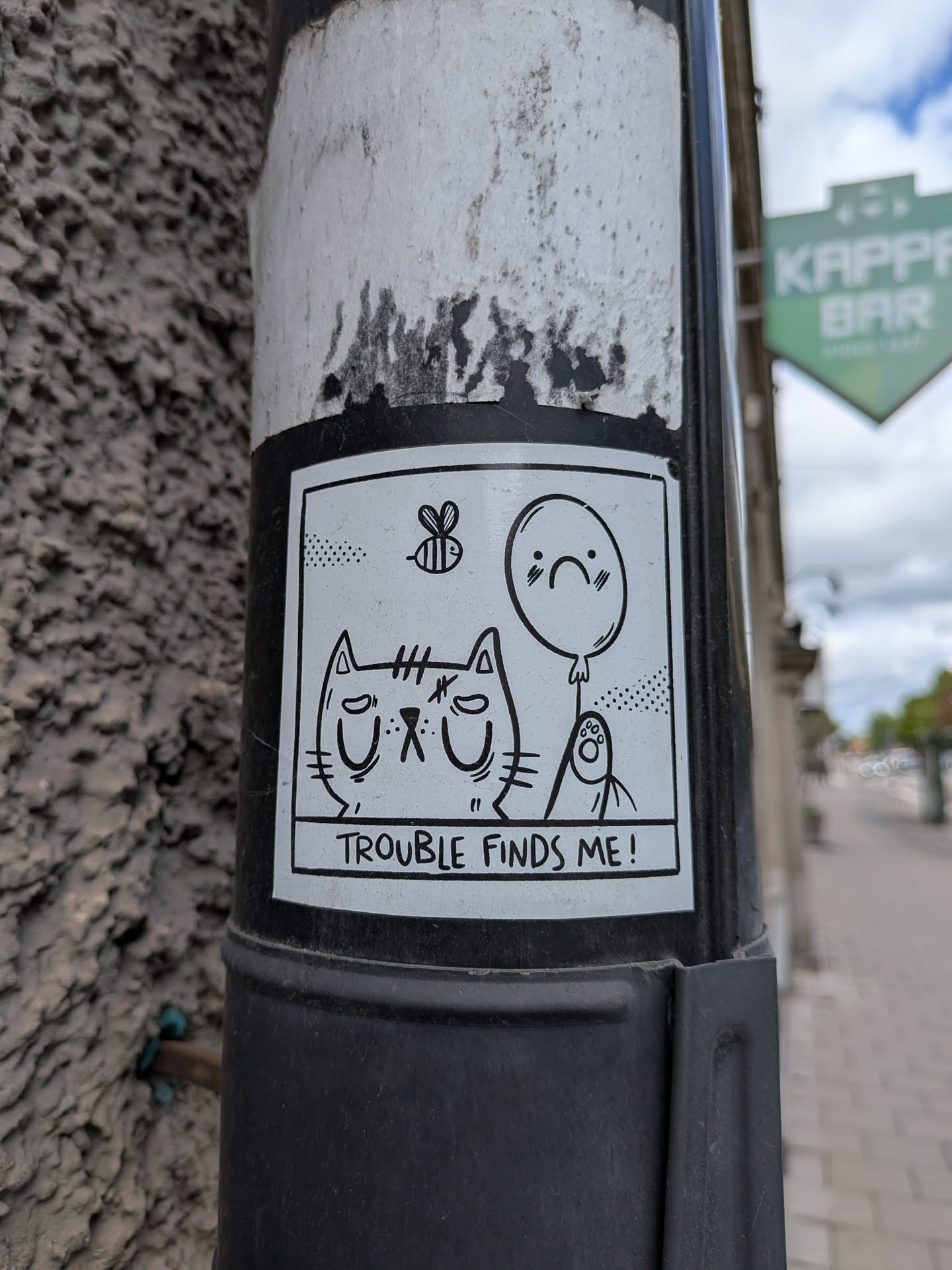 A rectangular, black and white cartoon sticker featuring a grumpy-looking cat with closed eyes and a downturned mouth. Above the cat, a small bee is depicted in flight. To the right of the cat, a balloon with a sad face floats, and below it, a small, round creature with a paw-like hand looks upwards. The entire scene is framed by a thick black border, and below the illustration, the text 'TROUBLE FINDS ME!' is written in a black box with white lettering.