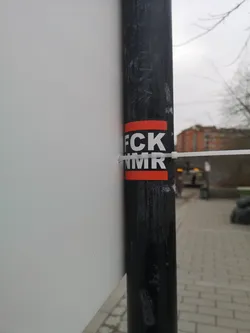 Street sticker The sticker is rectangular, featuring a black background with thick red borders at the top and bottom. White, sans-serif capital letters spell out 'FCK' on the upper black section and 'NMR' on the lower black section. The sticker is affixed to a textured black pole, with a white zip tie passing over its center, securing a white board or sign to the pole. The sticker itself appears slightly worn or creased in places.