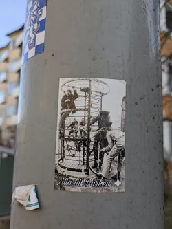 This sticker features a black and white image of people climbing a bicycle carousel, with the text "Alla till Parken" below the image. It's pasted on a light post, alongside another sticker.