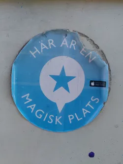 Street sticker A round blue sticker with a white speech bubble containing a blue star. The text around the circle says "HÄR ÄREN MAGISK PLATS" which translates to "Here is a magical place" in Swedish. There is also an App Store logo on the sticker. The sticker is slightly damaged around the edges.