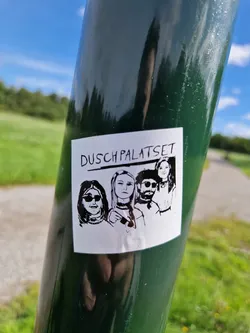 A black and white sticker featuring a stylized illustration of four individuals, likely a band or group. The text 'Dusch Palatset' is prominently displayed at the top. The sticker is affixed to a dark green pole outdoors.