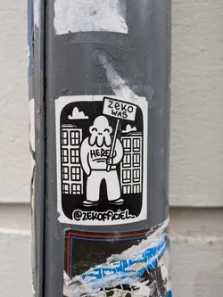 A black and white sticker featuring a stylized, bulbous, white character with three small eyes and a wide smile. The character holds a sign that reads 'ZEKO WAS HERE'. In the background, two blocky buildings with multiple windows are depicted against a solid black sky, with a small white cloud in the upper left. Below the character, the social media handle '@ZEKOFFICIEL' is visible. The sticker has a slightly worn appearance, consistent with being affixed to an outdoor pole.