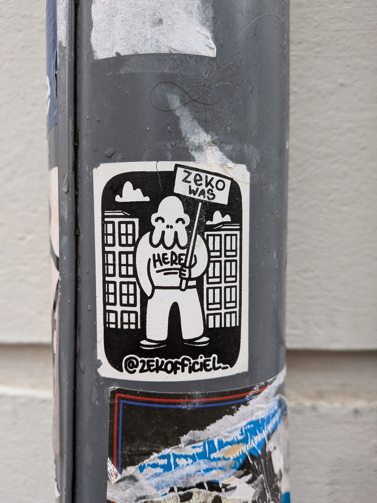 A black and white sticker featuring a stylized, bulbous, white character with three small eyes and a wide smile. The character holds a sign that reads 'ZEKO WAS HERE'. In the background, two blocky buildings with multiple windows are depicted against a solid black sky, with a small white cloud in the upper left. Below the character, the social media handle '@ZEKOFFICIEL' is visible. The sticker has a slightly worn appearance, consistent with being affixed to an outdoor pole.