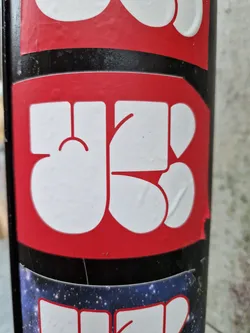 Street sticker Close-up of a red and white sticker with an abstract design. The sticker is affixed to a black surface, which is itself covered in other stickers. The main sticker features a stylized design of rounded, organic shapes in white against a red background. The style is reminiscent of modern or contemporary art, potentially street art or graffiti.