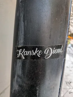 Street sticker A rectangular sticker featuring white cursive text on a solid black background. The sticker is applied to a textured, dark grey or black pole and shows signs of dust and minor wear, particularly around its edges. The text 'Kanske Diem!' is centrally placed and rendered in an elegant, flowing script, suggesting a humorous or ironic take on 'Carpe Diem'.